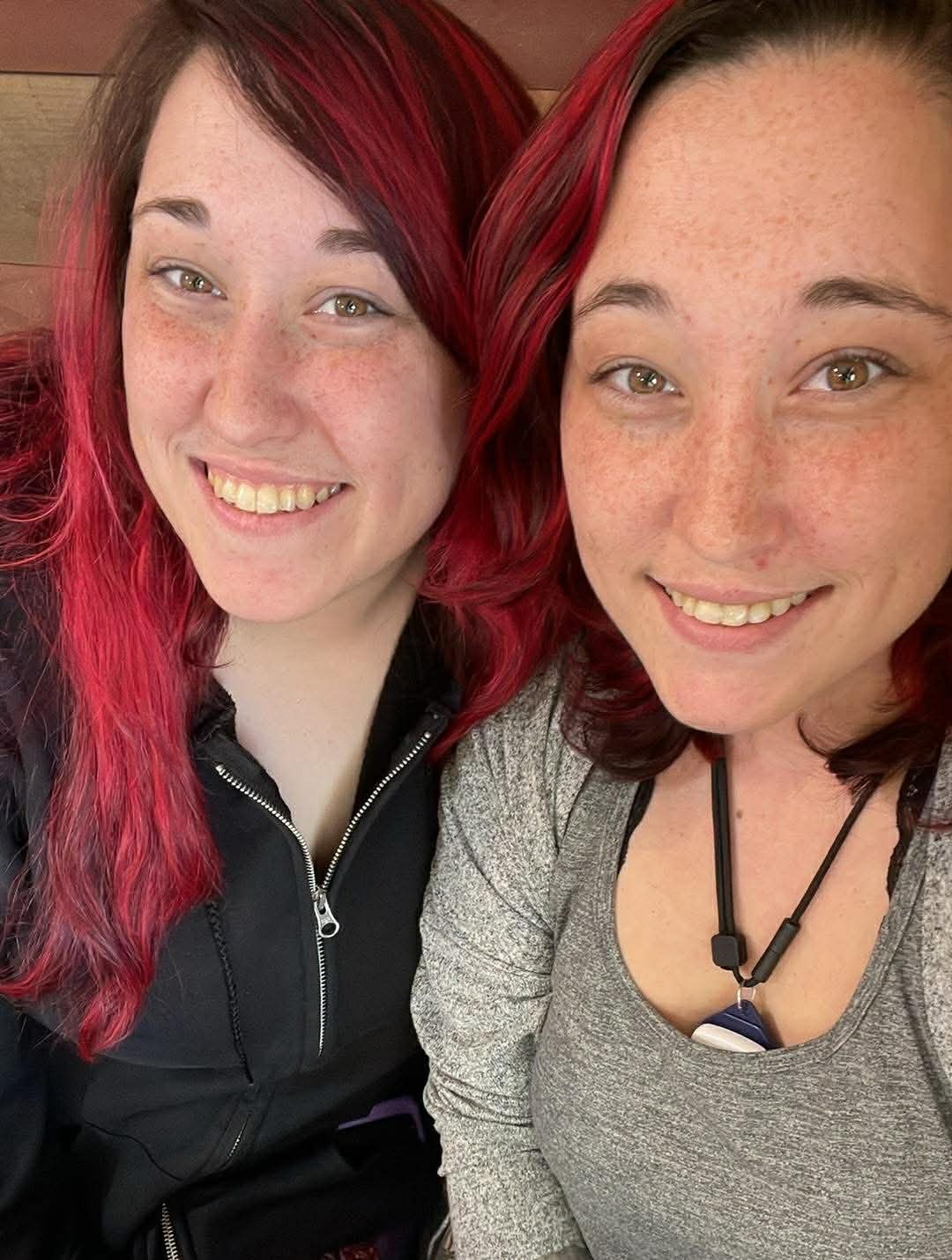 Two smiling twins with red hair wearing denim overalls, sitting close together and looking at the camera.